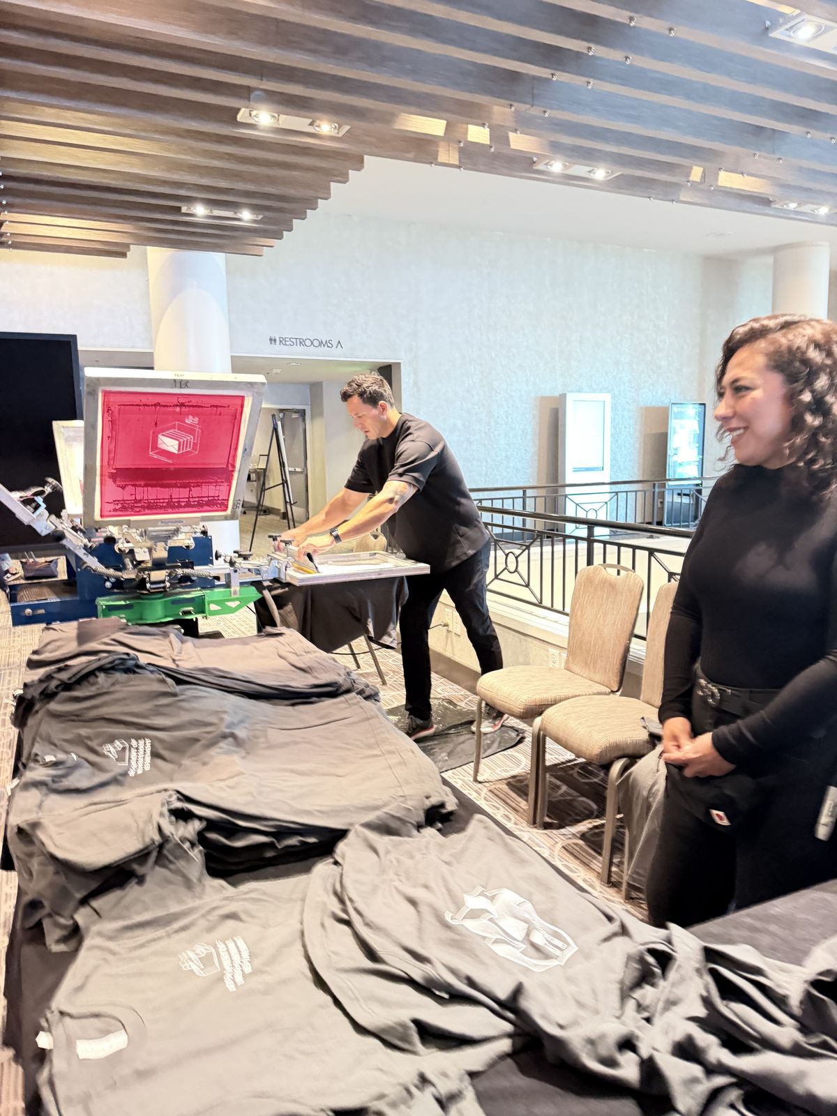 Stacks of freshly pressed tees piling up beside the press during a hotel conference event